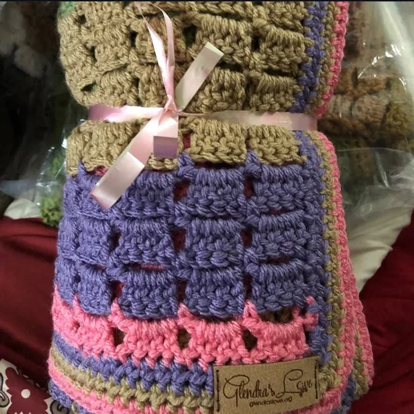 A crocheted blanket in tan, purple and pink tied with a pink ribbon, with a Glendra’s Love tag